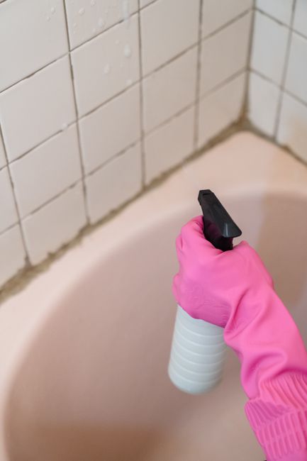 A close-up of a person wearing a bright pink rubber glove, holding a white spray bottle with a black nozzle, applying a cleaning solution to the edge of a bathtub. The background features a tiled bath