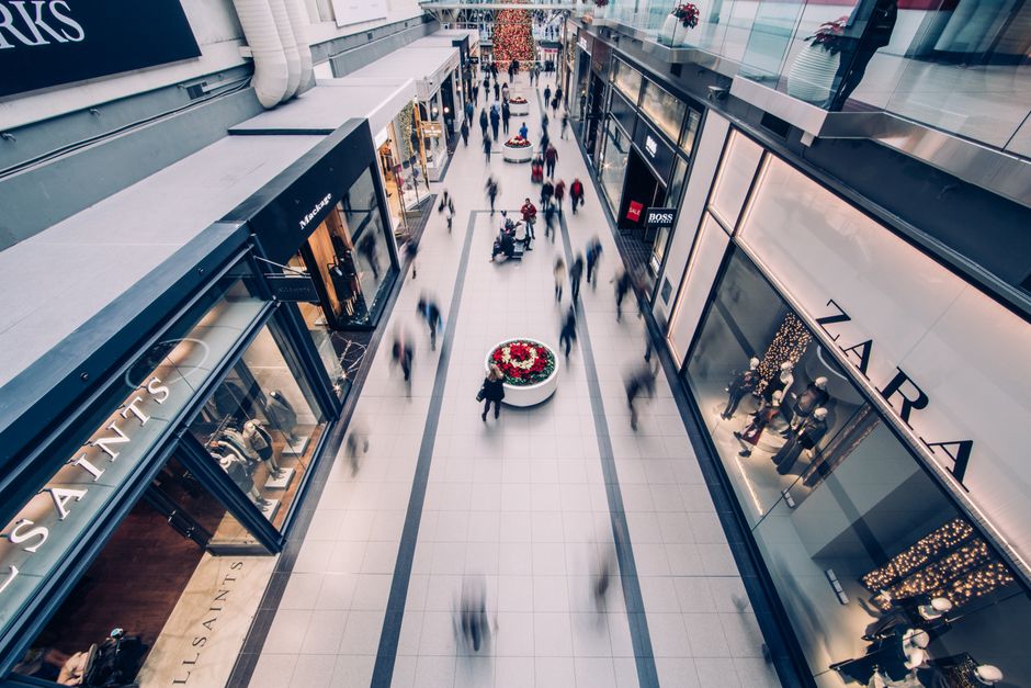 Exterior view of Brent Cross Shopping Centre showing a modern glass facade, metallic and concrete surfaces, with an outdoor commercial walkway featuring tiled flooring, potted plants, and a curved met